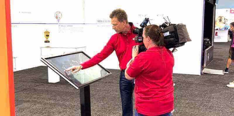 Camera operator filming man demonstrating interactive touchscreen kiosk at exhibition